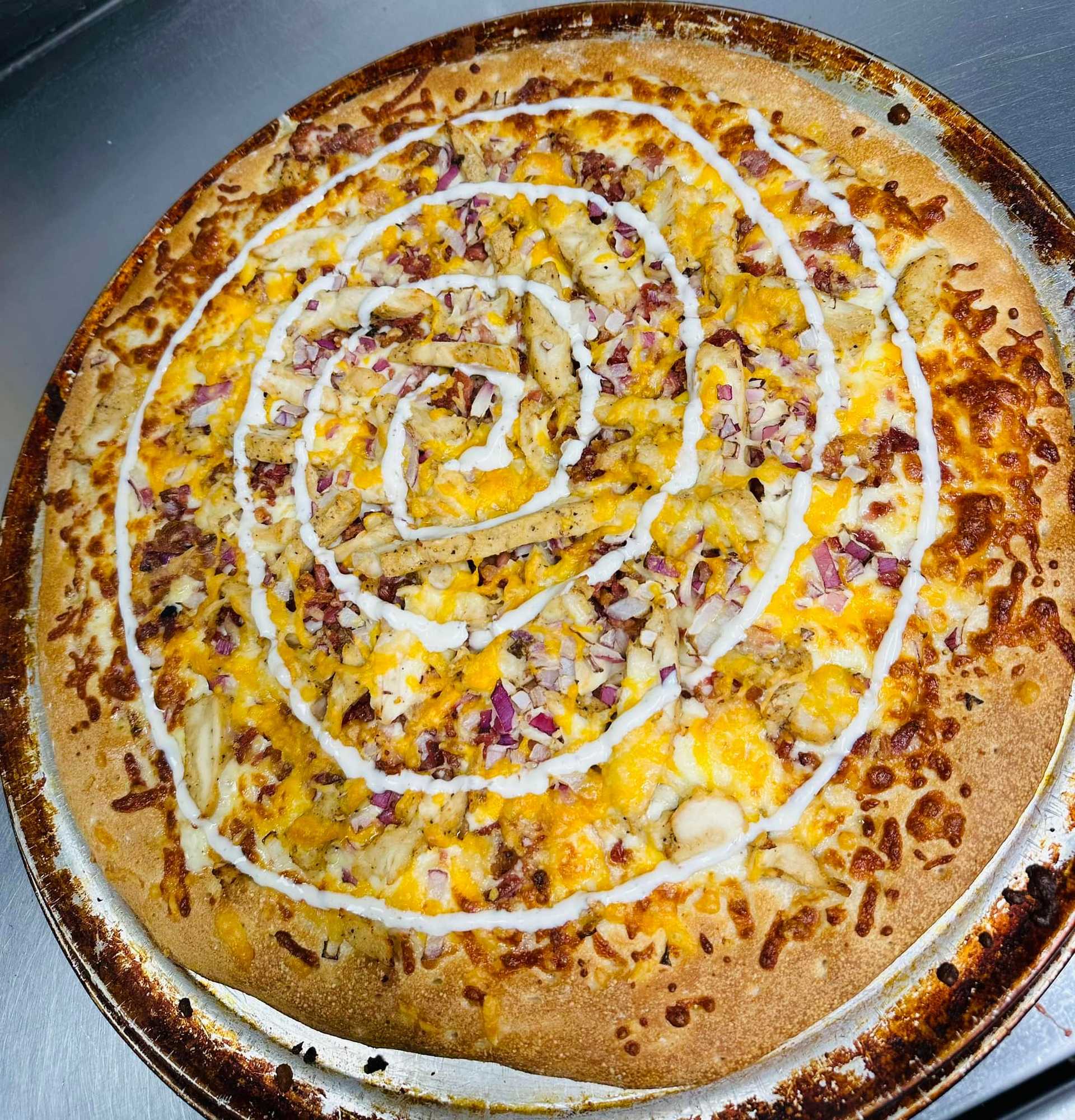 A loaded pizza topped with chicken, cheese, red onions, and a spiral of creamy sauce.