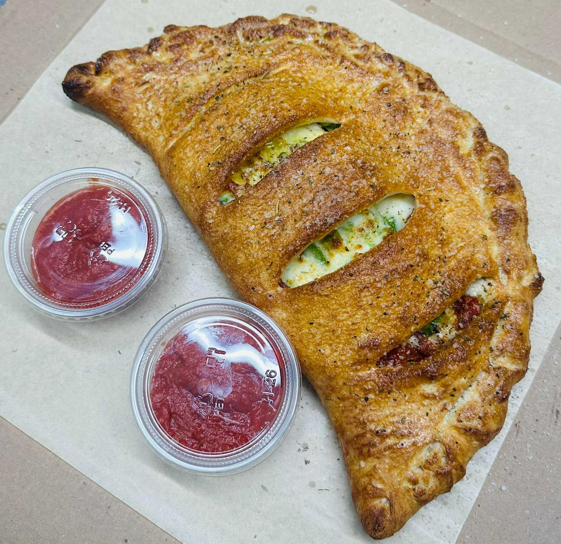 Golden-brown calzone with two cups of marinara sauce on a parchment paper, ready to serve.