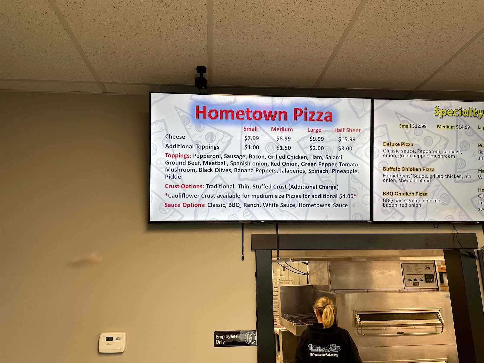 Digital menu board displaying options and prices for Hometown Pizza, with an employee in the background.